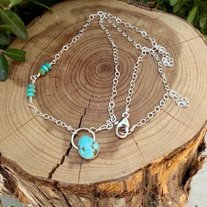May include: A silver chain necklace featuring a single teardrop-shaped turquoise pendant and a series of small, round turquoise beads along one side. The necklace includes a silver lobster claw clasp and a short extender chain for adjustable length.