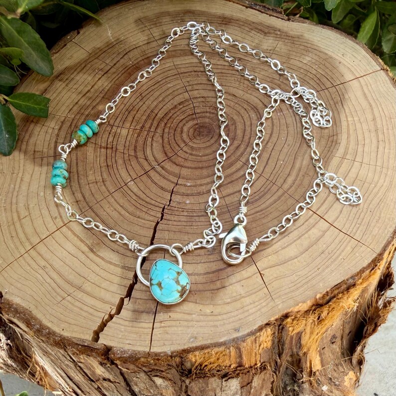 May include: A silver chain necklace featuring a single teardrop-shaped turquoise pendant and a series of small, round turquoise beads along one side. The necklace includes a silver lobster claw clasp and a short extender chain for adjustable length.