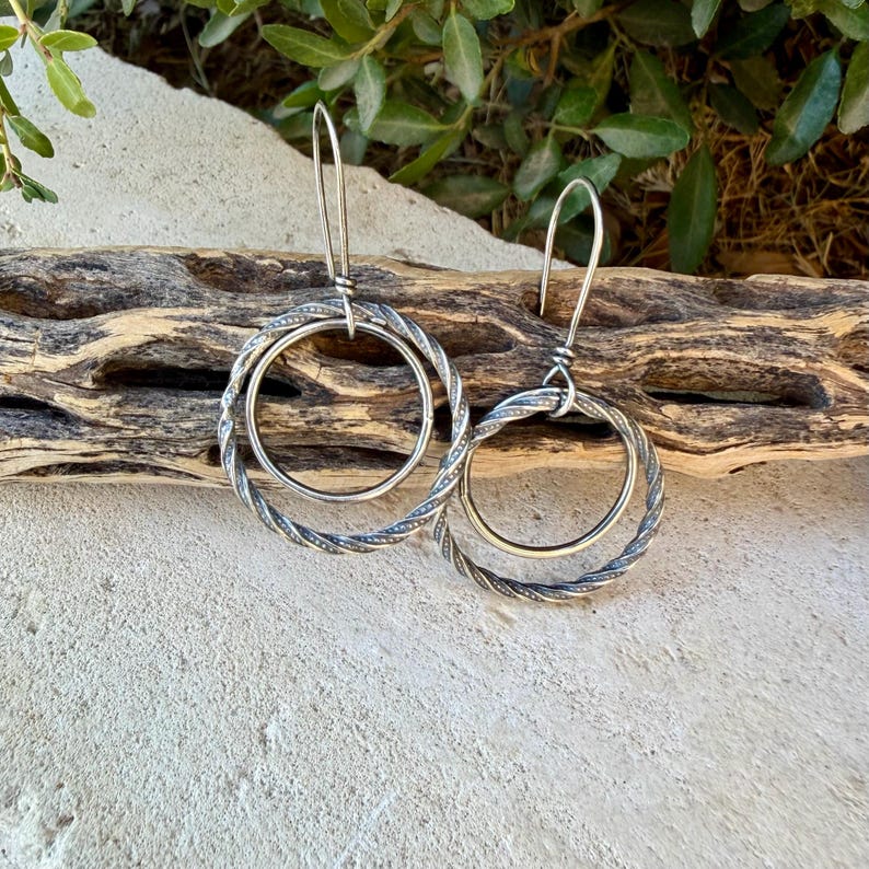 May include: A pair of silver-toned earrings featuring a double-hoop design. Each earring consists of a smaller smooth inner circle suspended within a larger, textured, twisted-wire outer circle, attached to a long, slender silver-toned ear wire.