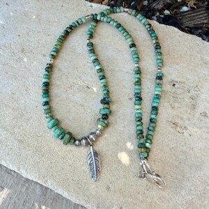 May include: A turquoise beaded necklace with a silver feather pendant. The necklace features small, round beads and larger, irregular-shaped beads. The feather pendant is detailed with etched lines. The necklace is secured with a silver clasp.