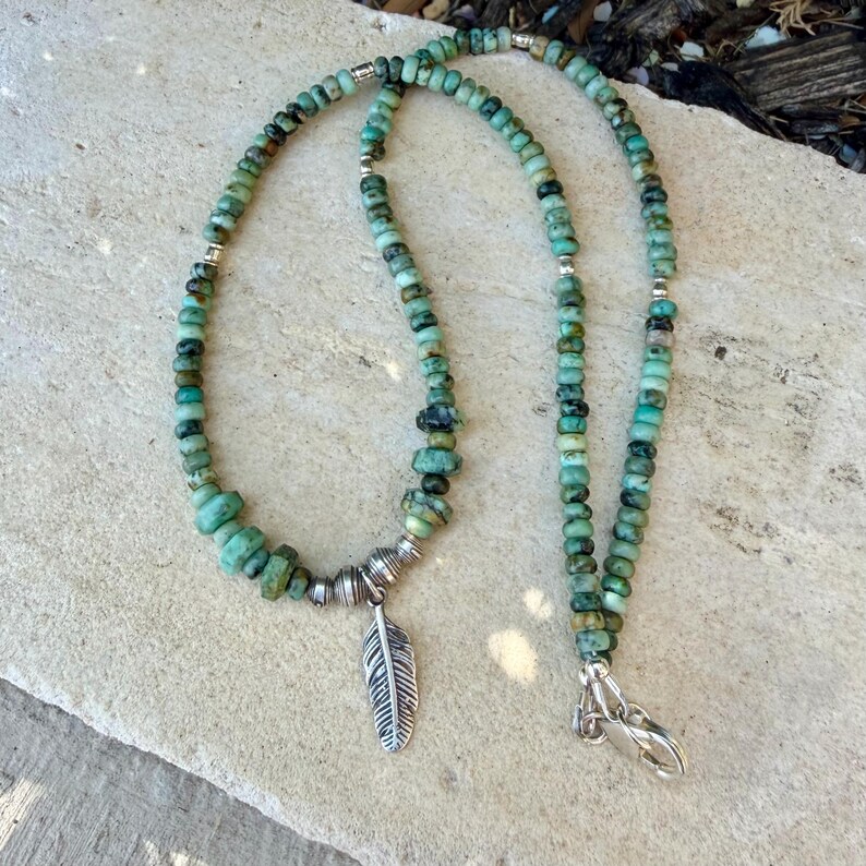 May include: A turquoise beaded necklace with a silver feather pendant. The necklace features small, round beads and larger, irregular-shaped beads. The feather pendant is detailed with etched lines. The necklace is secured with a silver clasp.