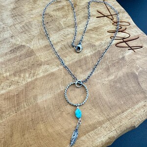 May include: Silver-toned necklace with a circular pendant featuring a turquoise bead and a feather charm. The necklace has a delicate chain and is displayed on a wooden surface.