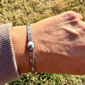 May include: A silver cuff bracelet featuring an oval, white and grey stone. The bracelet has an etched design along the sides. The bracelet is worn on a wrist.