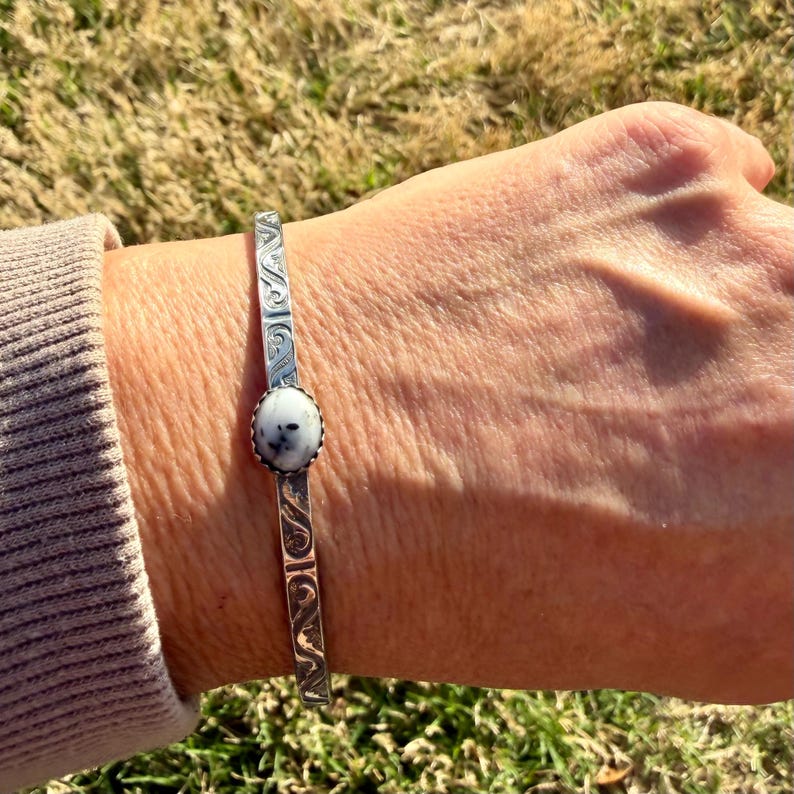 May include: A silver cuff bracelet featuring an oval, white and grey stone. The bracelet has an etched design along the sides. The bracelet is worn on a wrist.