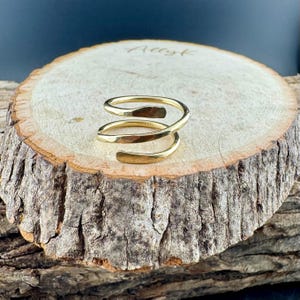 May include: A gold-colored spiral ring with a hammered texture. The ring is displayed on a rustic wooden surface, showcasing its unique design. The ring's open design allows for adjustable sizing.