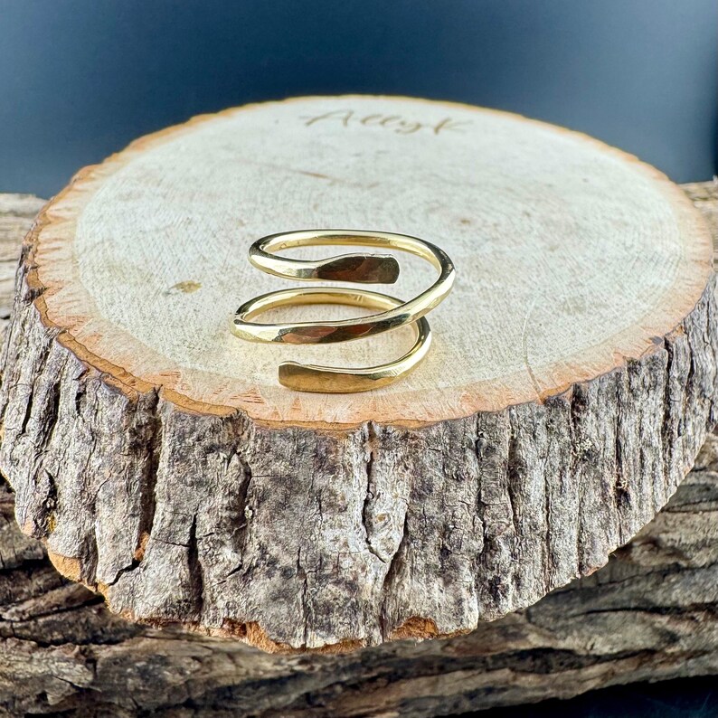 May include: A gold-colored spiral ring with a hammered texture. The ring is displayed on a rustic wooden surface, showcasing its unique design. The ring's open design allows for adjustable sizing.