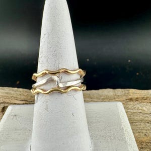 Puede incluir: Una pila de tres anillos, dos dorados y uno plateado, exhibidos en un soporte blanco en forma de cono. Los anillos tienen un dise&ntilde;o ondulado, creando un aspecto &uacute;nico y elegante. El fondo es oscuro, resaltando la joyer&iacute;a.