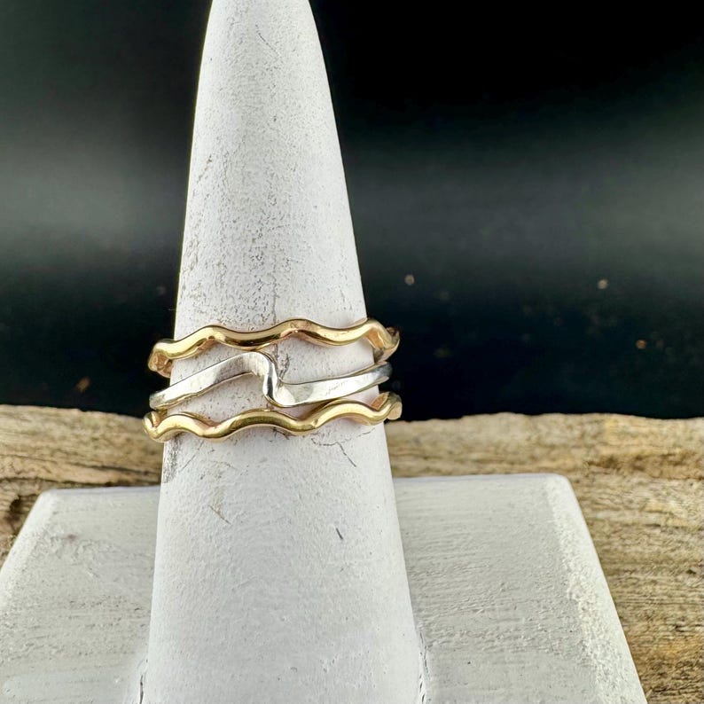 Puede incluir: Una pila de tres anillos, dos dorados y uno plateado, exhibidos en un soporte blanco en forma de cono. Los anillos tienen un dise&ntilde;o ondulado, creando un aspecto &uacute;nico y elegante. El fondo es oscuro, resaltando la joyer&iacute;a.