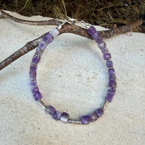Puede incluir: Una pulsera de cuentas con cubos de amatista morada facetada dispuestos en un patr&oacute;n circular. La pulsera incluye peque&ntilde;as cuentas espaciadoras de metal plateado con patrones intrincados y un cierre de mosquet&oacute;n plateado.