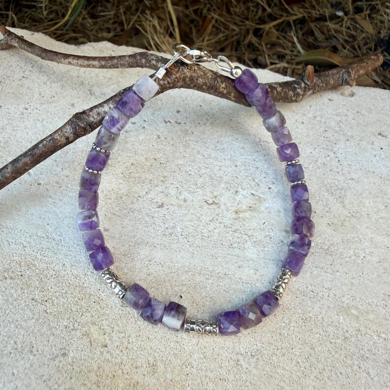 Puede incluir: Una pulsera de cuentas con cubos de amatista morada facetada dispuestos en un patr&oacute;n circular. La pulsera incluye peque&ntilde;as cuentas espaciadoras de metal plateado con patrones intrincados y un cierre de mosquet&oacute;n plateado.