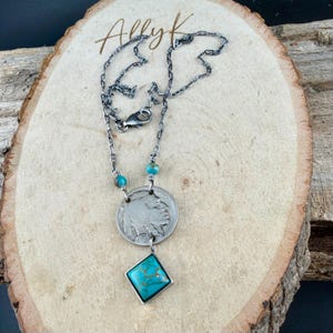 Op de afbeelding: Een zilveren ketting met een vintage munt hanger met een Native American profiel. De ketting bevat turquoise kralen en een vierkante turquoise stenen hanger. De ketting is donker zilverkleurig en wordt gepresenteerd op een houten oppervlak.