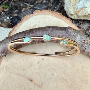 May include: A gold-colored cuff bracelet with three turquoise-colored stones. The bracelet has a double-band design and is displayed on a wooden surface. The stones are round and have a mottled appearance.