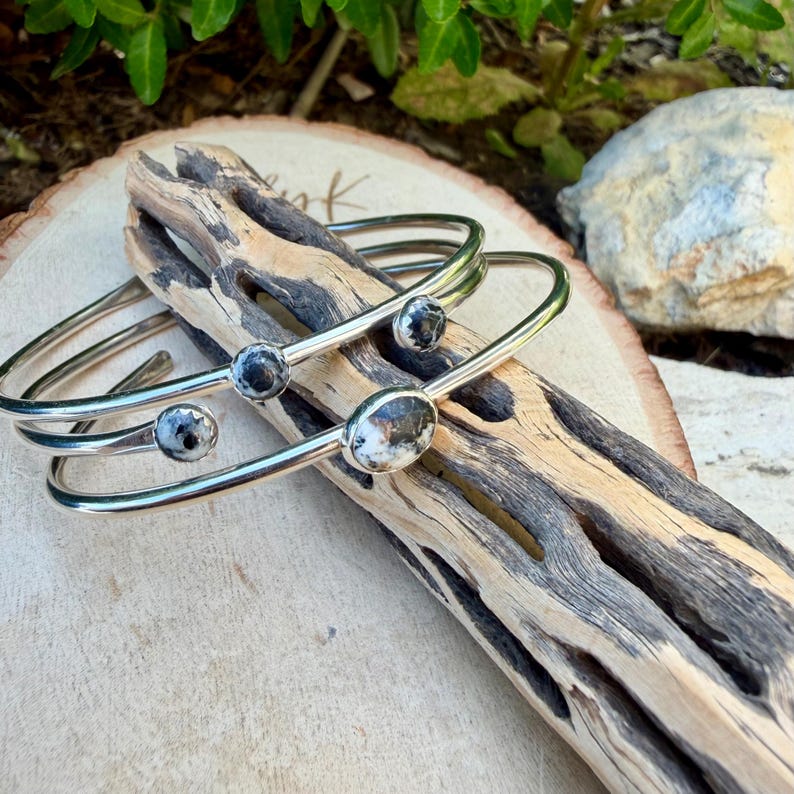 May include: Two silver-toned metal bangles with oval and round black and white speckled stones. The bracelets are displayed on a piece of driftwood, showcasing their design. The background includes greenery and a light-colored stone.