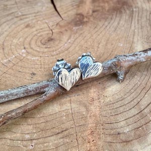 May include: A pair of silver heart-shaped stud earrings featuring a textured, hammered surface finish. Each earring includes a standard post back mechanism, showcasing a reflective metallic sheen and a distinct, irregular pattern across the front.