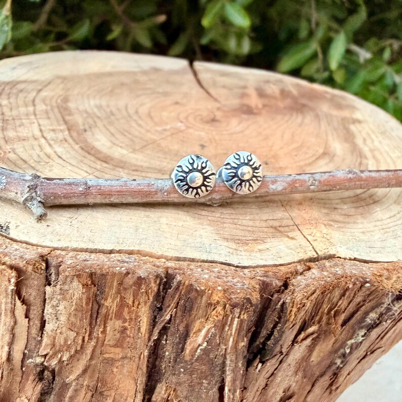 May include: A pair of circular silver stud earrings featuring a raised sun design with radiating rays and a smooth central dome. The earrings are positioned side by side on a thin, light brown wooden twig.