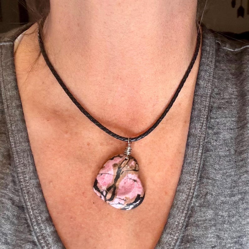 May include: A pink and black stone pendant necklace. The pendant is a unique, irregularly shaped stone with pink and black patterns. It hangs from a black, braided cord necklace. The necklace is worn around the neck.