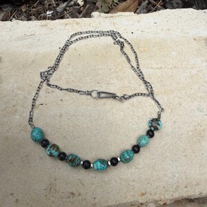 May include: A silver chain necklace featuring turquoise and black beads. The necklace has a simple clasp and is displayed on a neutral-colored surface. The beads are interspersed along the chain, creating a unique jewelry piece.