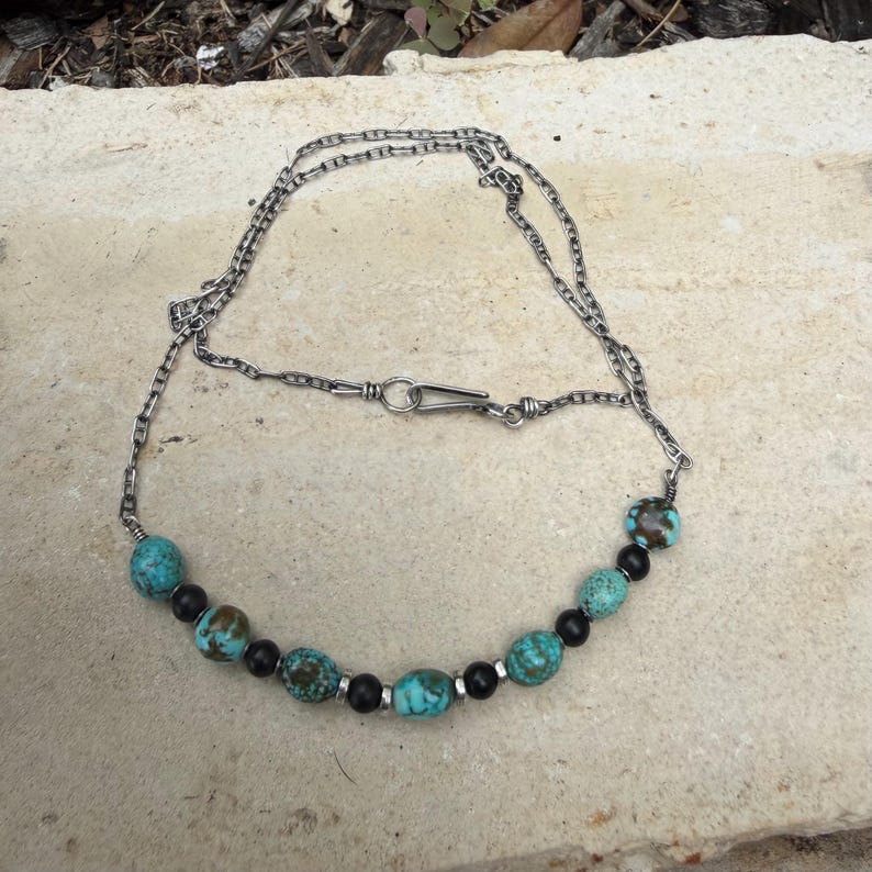 May include: A silver chain necklace featuring turquoise and black beads. The necklace has a simple clasp and is displayed on a neutral-colored surface. The beads are interspersed along the chain, creating a unique jewelry piece.