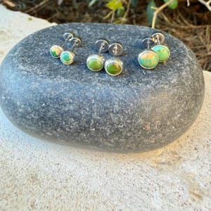 May include: Three pairs of stud earrings featuring round, polished green turquoise stones set in silver-toned metal settings. Each earring has a post back and a circular stone face with natural veining patterns in shades of lime and forest green.