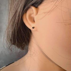 May include: A close-up of a person's ear with a small, black stud earring. The earring is a simple, round design. The person has dark hair pulled back, and the skin tone is fair.