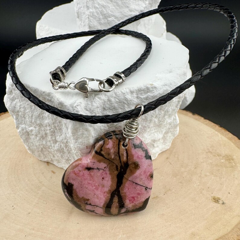 May include: A heart-shaped pendant necklace. The pendant is a mix of pink and brown tones, with a silver wire wrap. The necklace has a black braided leather cord with a silver-colored clasp.