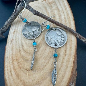 May include: Silver earrings with vintage buffalo nickel coins, turquoise beads, and feather charms. The earrings have a rustic, Native American-inspired design, with a hook closure. The coins are engraved with the words "FIVE CENTS".