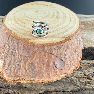 May include: A silver-colored ear cuff with a turquoise stone set in the center. The cuff has a layered design with multiple bands wrapping around the ear. The jewelry is displayed on a wooden surface.