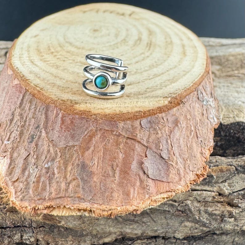 May include: A silver-colored ear cuff with a turquoise stone set in the center. The cuff has a layered design with multiple bands wrapping around the ear. The jewelry is displayed on a wooden surface.