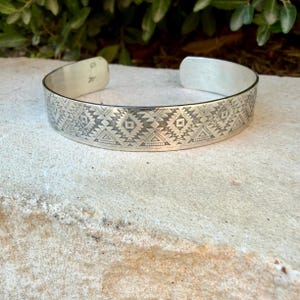 May include: A silver cuff bracelet featuring a repeating geometric pattern of triangles, diamonds, and small dots engraved across the outer surface. The metal has a polished finish, and the interior band shows a small hallmark stamp near the opening.