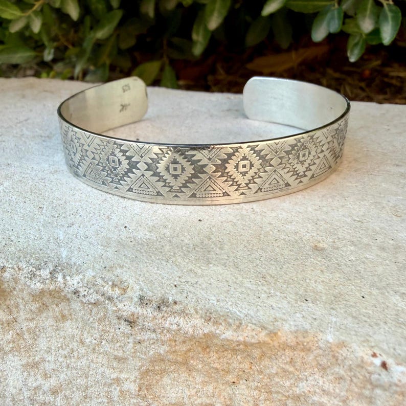 May include: A silver cuff bracelet featuring a repeating geometric pattern of triangles, diamonds, and small dots engraved across the outer surface. The metal has a polished finish, and the interior band shows a small hallmark stamp near the opening.