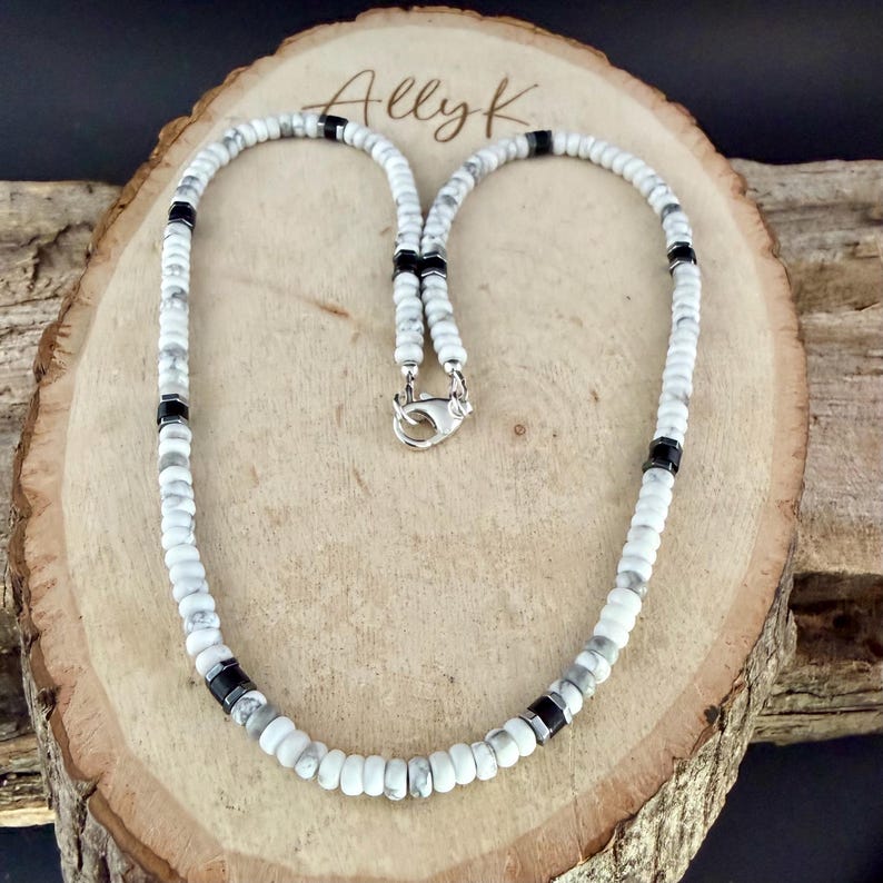 May include: White and gray beaded necklace with black accents and a silver clasp. The necklace is displayed on a wooden surface with the name "Allyk" engraved on it. The beads are small and round.