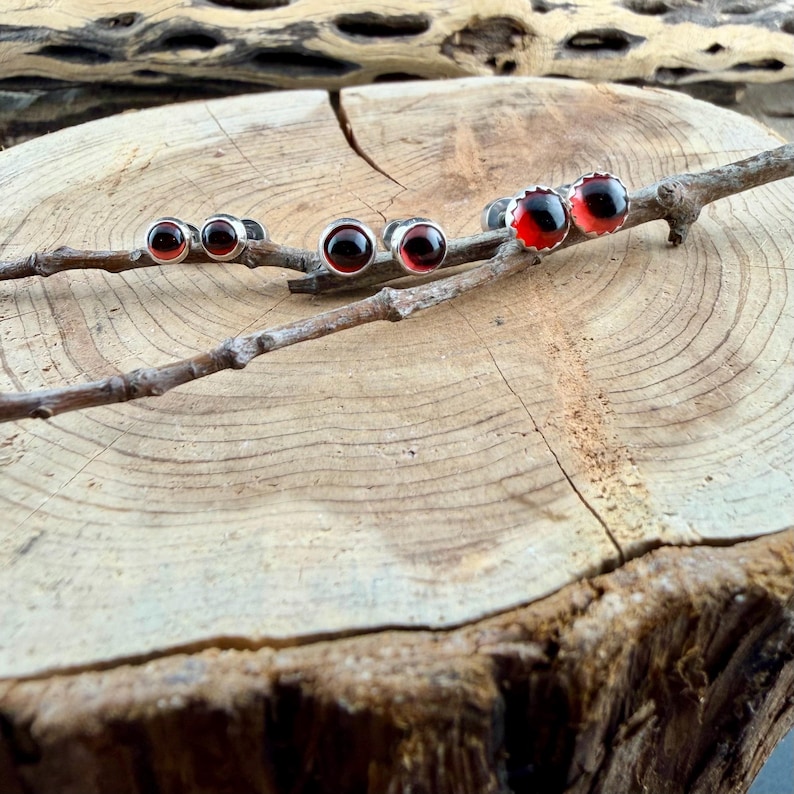 May include: Three pairs of stud earrings featuring round, deep red cabochon stones set in polished silver-toned metal settings. The earrings are arranged in a row along a thin, brown, textured wooden twig.