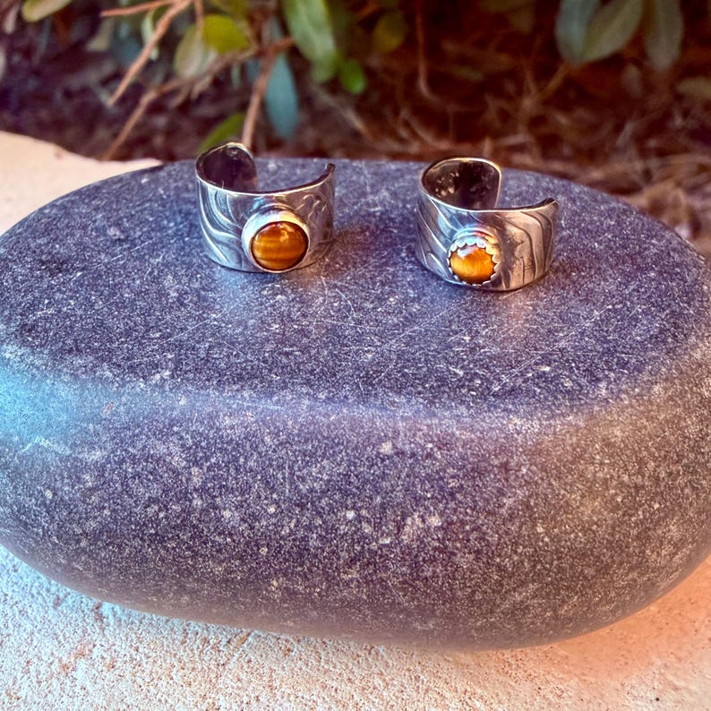 K&ouml;nnte beinhalten: Zwei offene Ringe aus silberfarbenem Metall mit einer strukturierten, wellenf&ouml;rmigen Oberfl&auml;che. Jeder Ring ist mit einem einzelnen, runden, polierten Tigerauge-Edelstein besetzt, der in einer glatten Fassung sitzt.