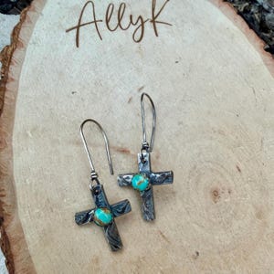 May include: A pair of silver-toned cross earrings with turquoise-colored stones at the center. The earrings have a textured design and long, curved ear wires. The image also includes the text "Allyk" in a handwritten font.