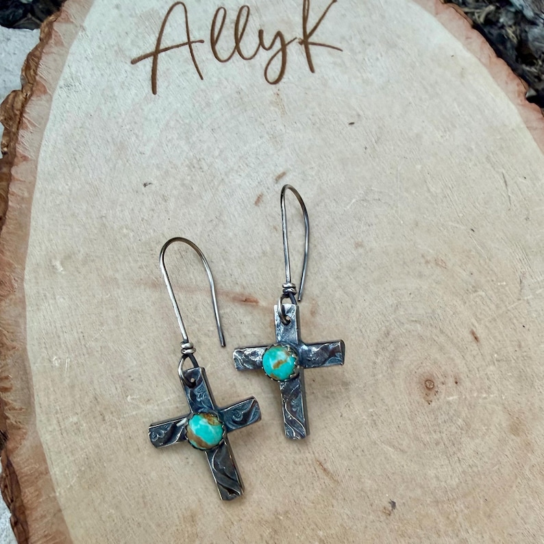 May include: A pair of silver-toned cross earrings with turquoise-colored stones at the center. The earrings have a textured design and long, curved ear wires. The image also includes the text "Allyk" in a handwritten font.