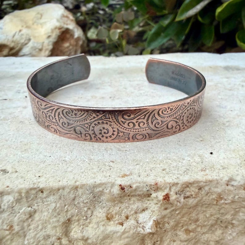 Puede incluir: Brazalete de cobre con un intrincado dise&ntilde;o en espiral. El brazalete abierto tiene un interior oscuro y un dise&ntilde;o detallado en relieve en el exterior. El metal tiene un tono marr&oacute;n rojizo c&aacute;lido, y el dise&ntilde;o es visible en la superficie.