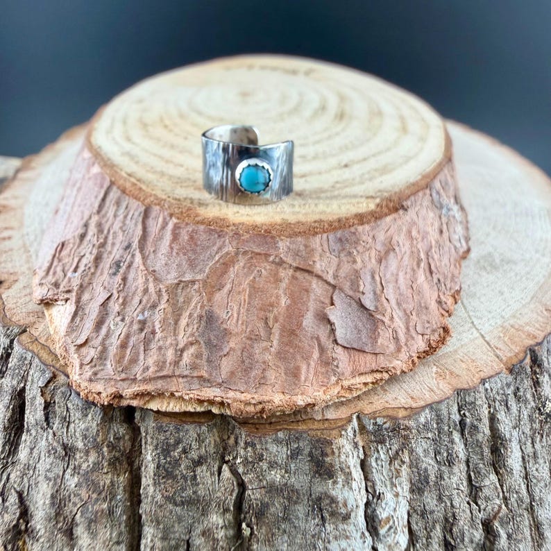 May include: A silver-toned ring with a textured band and a round, turquoise-colored stone. The ring is set on a rustic wooden surface, showcasing its handcrafted appearance. The ring's design is simple yet elegant, suitable for everyday wear.