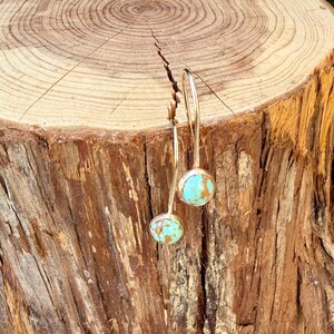 May include: A pair of drop earrings featuring round, polished turquoise stones with brown matrix veining. Each stone is set in a simple silver-toned metal bezel, suspended from a curved silver-toned wire hook that hangs vertically from the top.