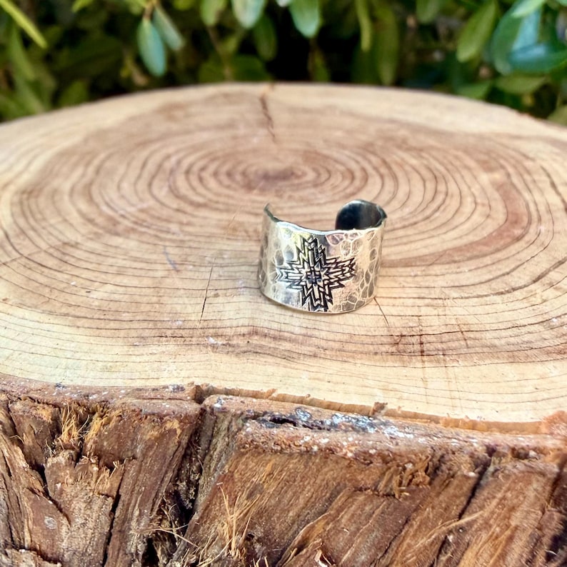 Puede incluir: Un anillo de metal color plata con dise&ntilde;o de banda abierta, textura martillada y un patr&oacute;n de estrella geom&eacute;trica grabado en el centro. El anillo tiene un acabado pulido y una forma ancha y curva que se envuelve parcialmente alrededor del dedo.
