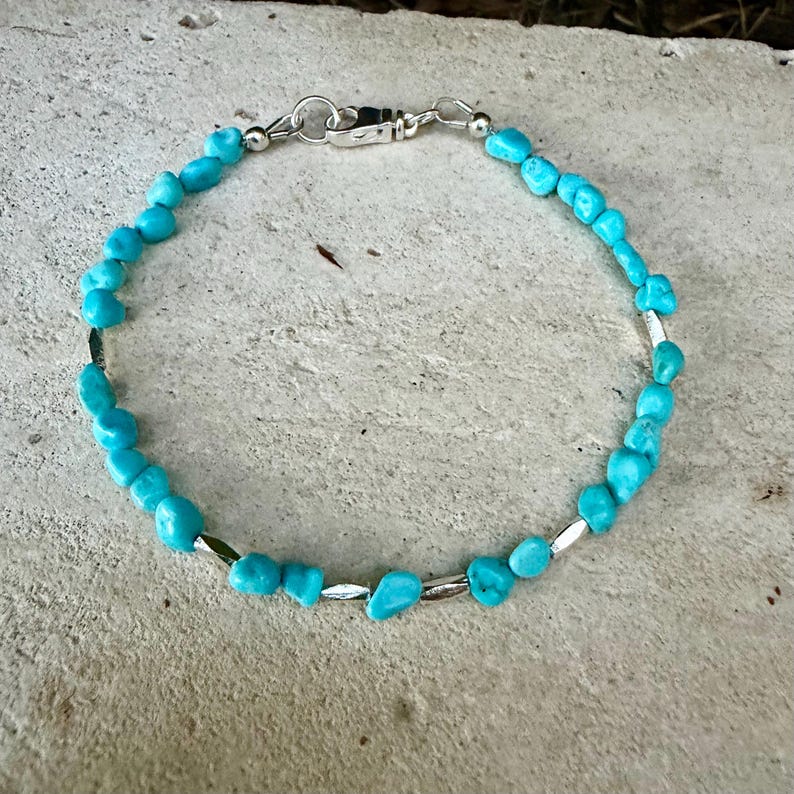 May include: A bracelet featuring small, irregular, bright turquoise-colored stone beads interspersed with thin, metallic silver-toned cylindrical spacers. The piece is secured with a silver-toned metal lobster claw clasp.