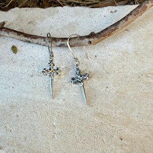 May include: A pair of silver-toned drop earrings featuring a sword-shaped pendant with an ornate cross-guard design. Each earring hangs from a simple curved wire hook, showcasing intricate metalwork details on the hilt and a pointed blade tip.