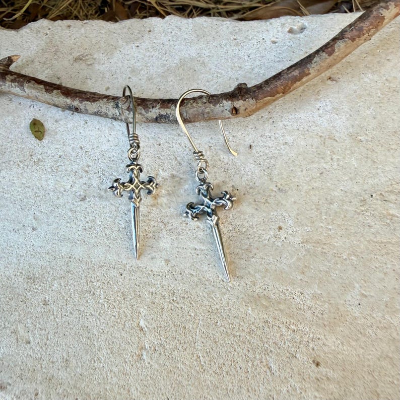 May include: A pair of silver-toned drop earrings featuring a sword-shaped pendant with an ornate cross-guard design. Each earring hangs from a simple curved wire hook, showcasing intricate metalwork details on the hilt and a pointed blade tip.