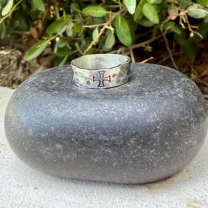 May include: A silver-toned metal ring with a hammered, textured surface and a prominent black cross symbol engraved on the front. The band has a wide, flat profile and a polished finish, showcasing a distinct contrast between the dark cross and metal.