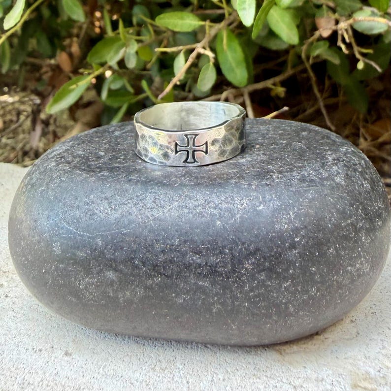 May include: A silver-toned metal ring with a hammered, textured surface and a prominent black cross symbol engraved on the front. The band has a wide, flat profile and a polished finish, showcasing a distinct contrast between the dark cross and metal.