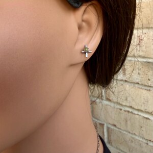 K&ouml;nnte beinhalten: Ein silberner Ohrstecker in Kreuzform mit einem kleinen, runden, hellgr&uuml;nen Stein in der Mitte. Der Ohrring wird am Ohr einer Person getragen. Der Ohrring hat ein schlichtes, elegantes Design.