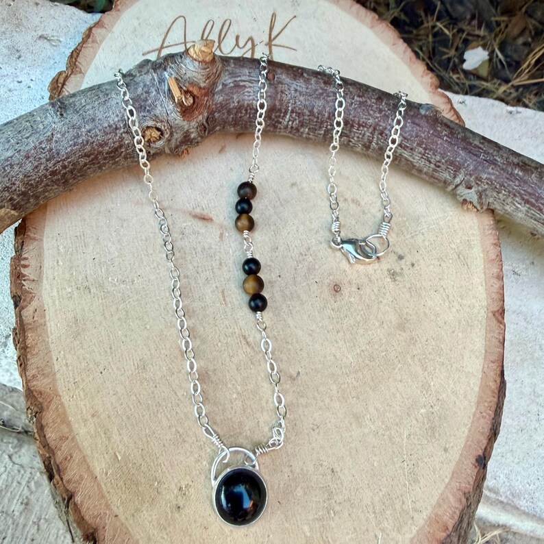 May include: A silver-toned chain necklace featuring a circular black pendant at the base and a series of alternating black and brown spherical beads integrated into one side of the chain, secured with a lobster claw clasp closure.