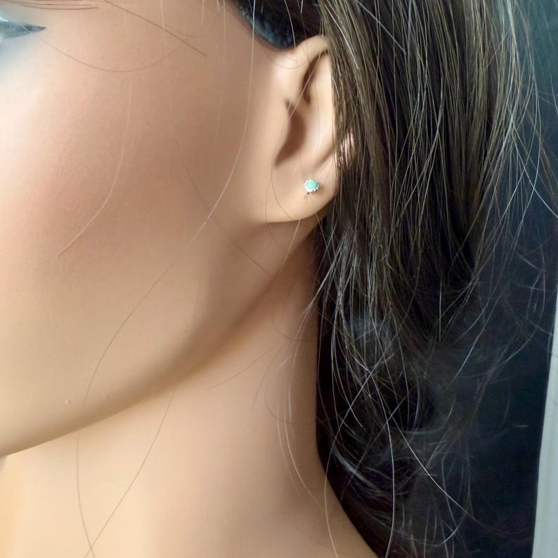 May include: A close-up of a person's ear with a small, round stud earring. The earring has a light blue center stone with a silver-colored setting. The person has brown hair.