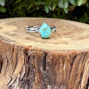 Puede incluir: Un anillo de plata con una piedra turquesa en forma de l&aacute;grima con vetas de matriz marr&oacute;n. La banda est&aacute; construida con m&uacute;ltiples hilos de plata trenzados que convergen para sostener la piedra central en un engaste de metal liso y pulido.