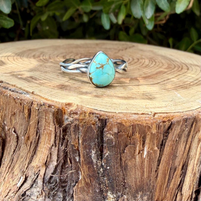 Puede incluir: Un anillo de plata con una piedra turquesa en forma de l&aacute;grima con vetas de matriz marr&oacute;n. La banda est&aacute; construida con m&uacute;ltiples hilos de plata trenzados que convergen para sostener la piedra central en un engaste de metal liso y pulido.