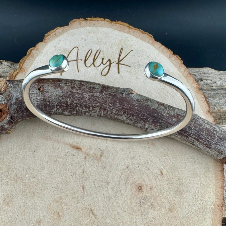 K&ouml;nnte beinhalten: Ein silbernes Armreif-Armband mit T&uuml;rkissteinen an jedem Ende. Das Armband wird auf einer Holzoberfl&auml;che mit dem handgeschriebenen Text "Ally K" pr&auml;sentiert. Die T&uuml;rkissteine sind rund und in einer silbernen L&uuml;nette gefasst.